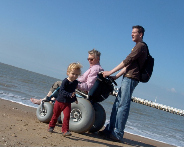 image of beach woman using a beach wheelchair, man standing and small child running on sandy beach and sea 
