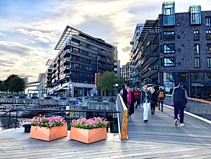 Oslo waterfront boardwalk with people crossing pedestrian bridge, pink flowers in wooden planters and modern apartment buildings with balconies in the background.