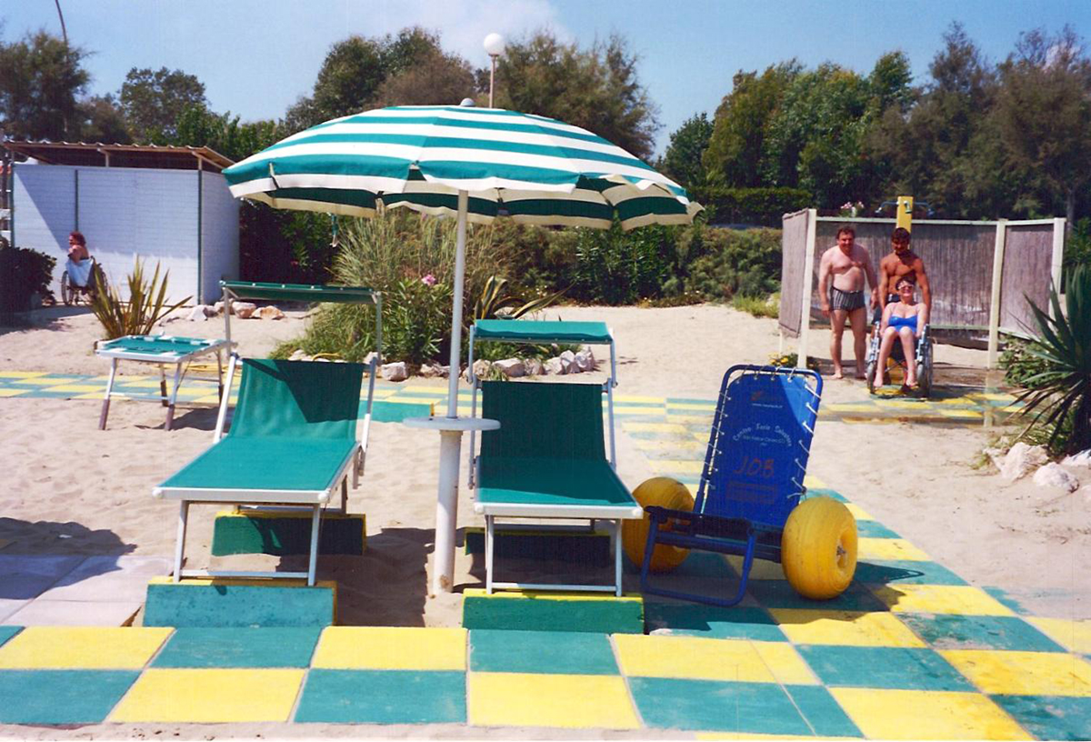 Two adapted sunbeds on a paved beach with a blue and yellow beach wheelchair next to them under an umbrella