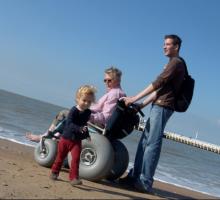 image of beach woman using a beach wheelchair, man standing and small child running on sandy beach and sea 